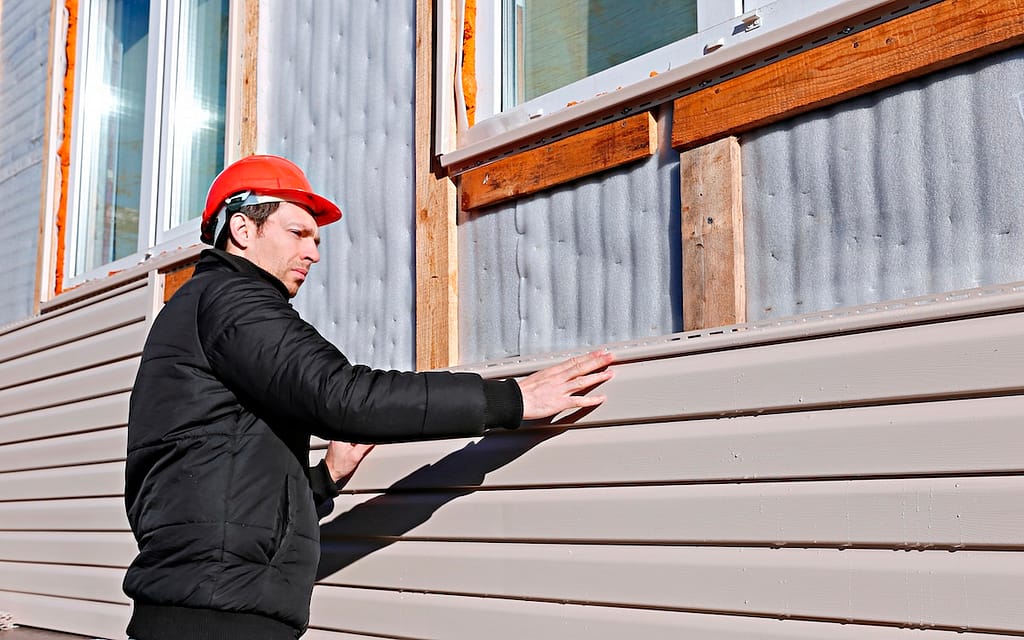 A worker installs panels beige vinyl siding on the facade of the house