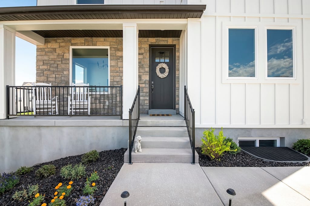 Facade of a house with stone veneer and white board and batten sidings. Entrance of a house with plants at the front of the basement window and porch with chairs and black front door with wreath.