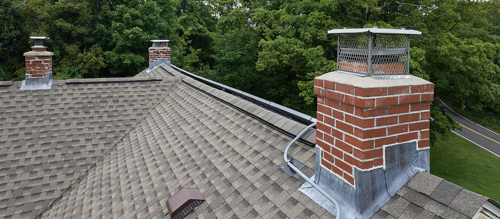 Multiple brick chimneys with metal flashing on a shingle roof surrounded by trees