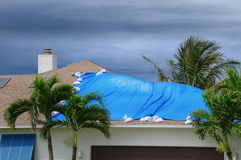 Storm damaged roof on house with a blue plastic tarp over hole in the shingles and rooftop.