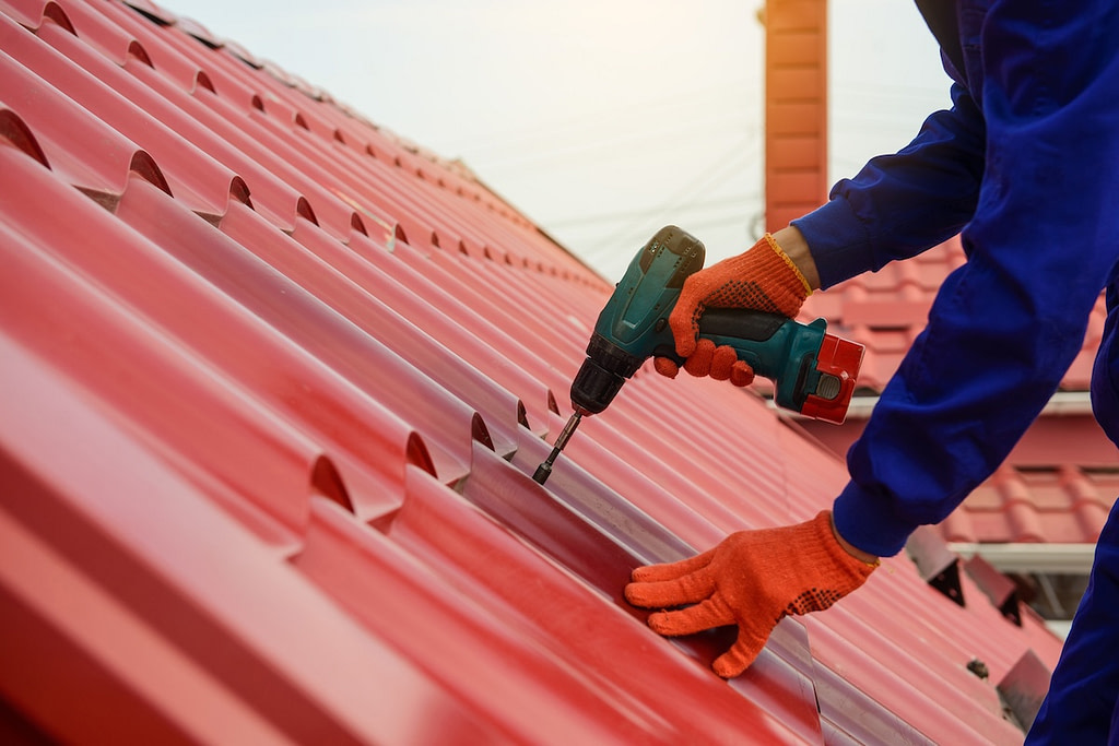 Close up hands in protective gloves of young man worker who fix a metal tile roof with screwdriver.