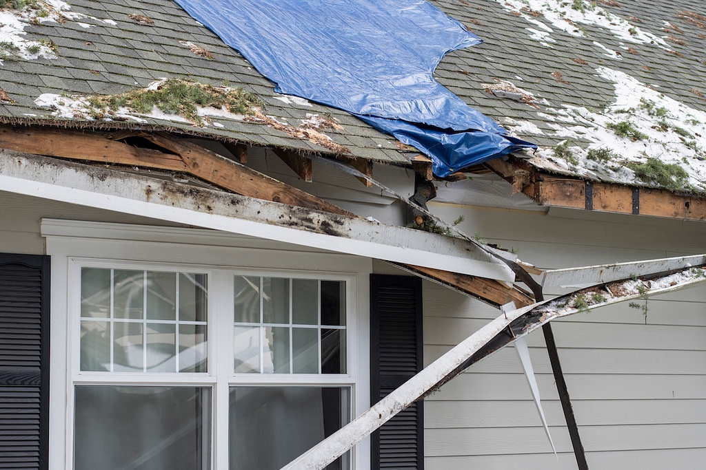 Residential house crushed by fallen trees and tree limbs during severe winter storm with strong winds. Tarp is placed on the damaged rooftop area as a temporary measure before proper roof repairs.