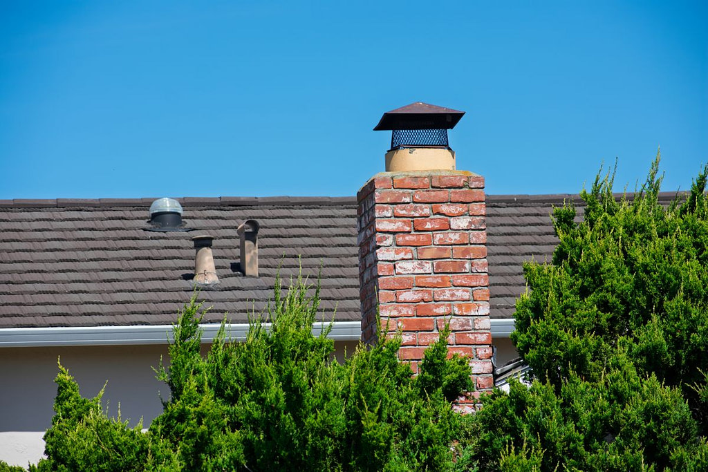 Red brick chimney with a black cap rises above a shingled roof, framed by green shrubs