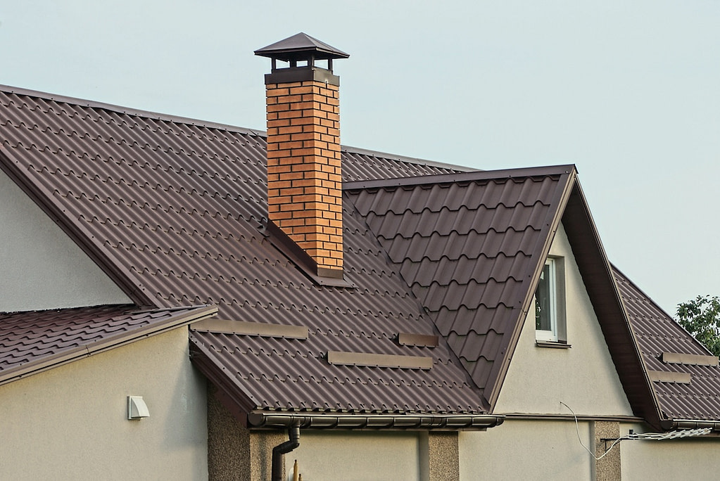 one large red brick chimney on a brown tiled roof of a private gray house with a window against the sky