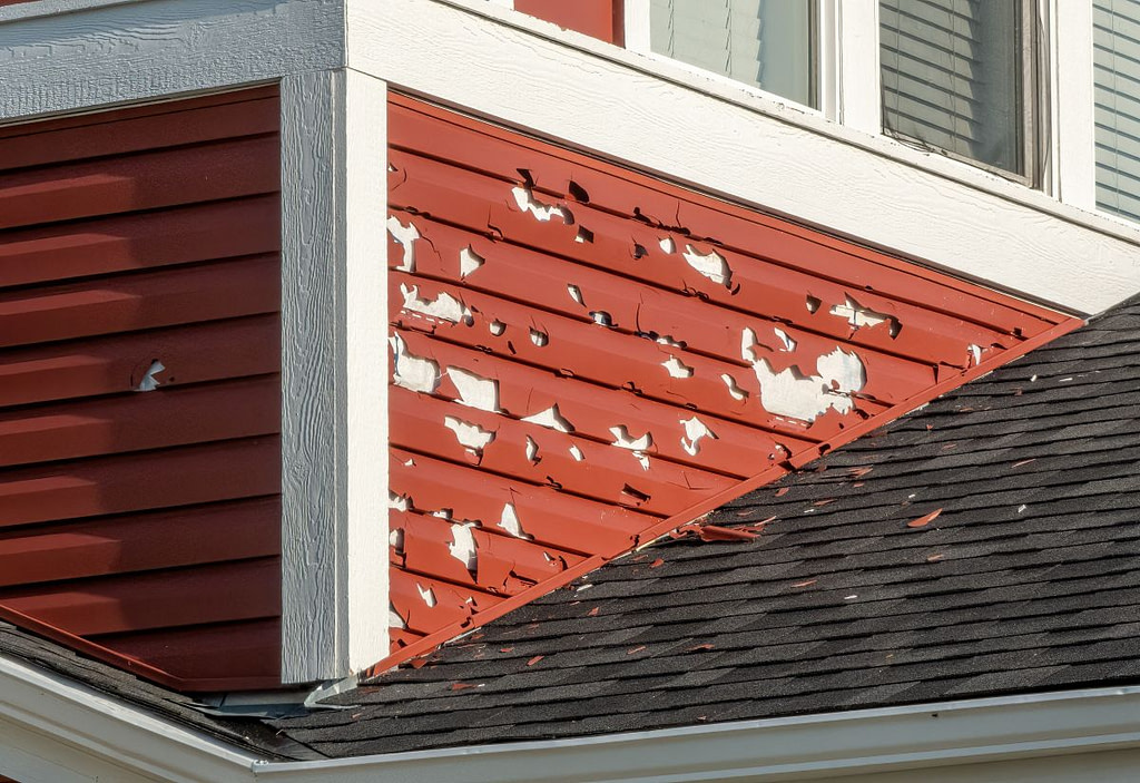 A close-up of a building's exterior shows red siding with extensive hail damage, exposing the underlying white surface