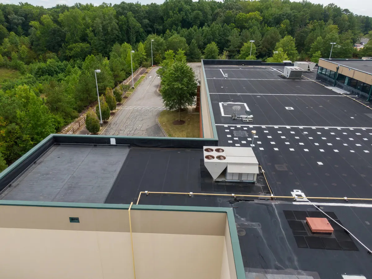 Large flat commercial roof with black rubber roofing and HVAC units, surrounded by trees and parking area