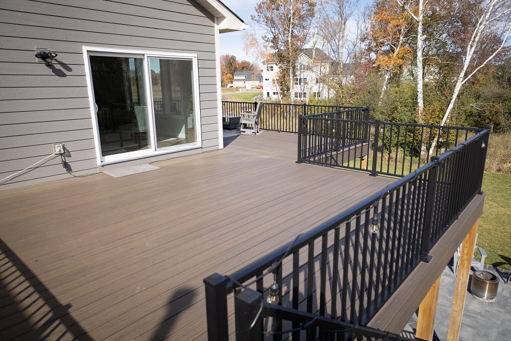 A spacious wooden deck with black metal railings extends from a house, featuring sliding glass doors. Surrounded by autumn trees, the deck overlooks a suburban neighborhood with green grass and a few houses in the distance.