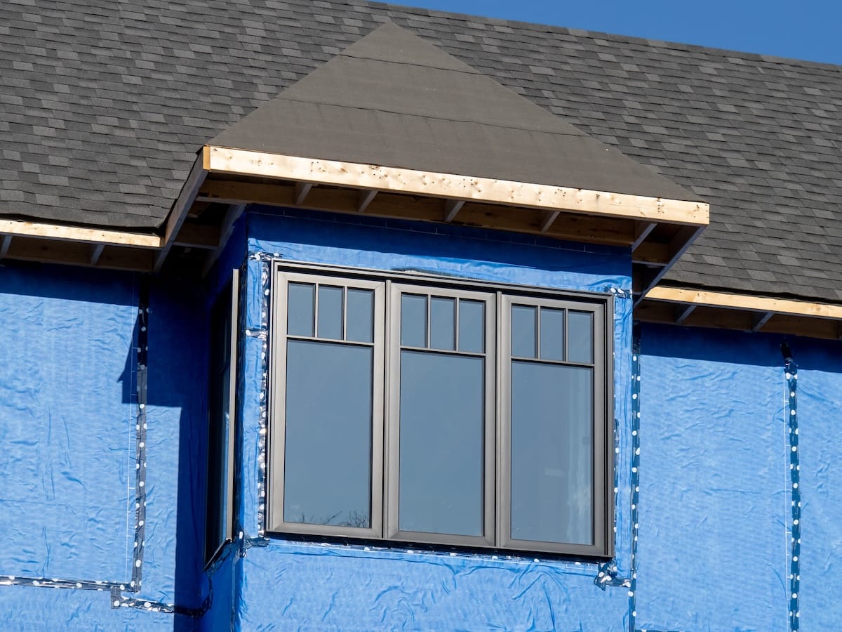 A house under construction features a gray-framed window and a triangular gable. The exterior is covered in blue weather-resistant house wrap