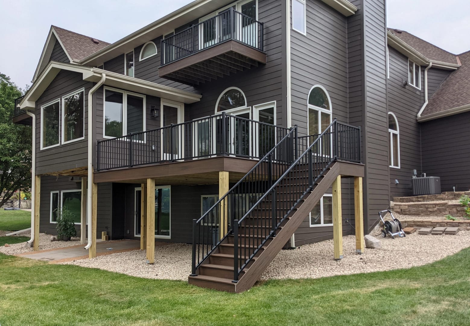 A modern brown two-story house with large windows, two elevated decks, black railings, and a staircase leading to the grassy backyard. The area under the deck is covered with stones and wooden support posts.