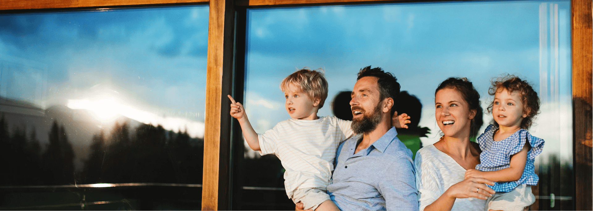 A family of four stands in front of a window during sunset. The father holds a young boy, who is pointing, and the mother holds a young girl. They all smile and look in the same direction. The sky reflects in the glass behind them.
