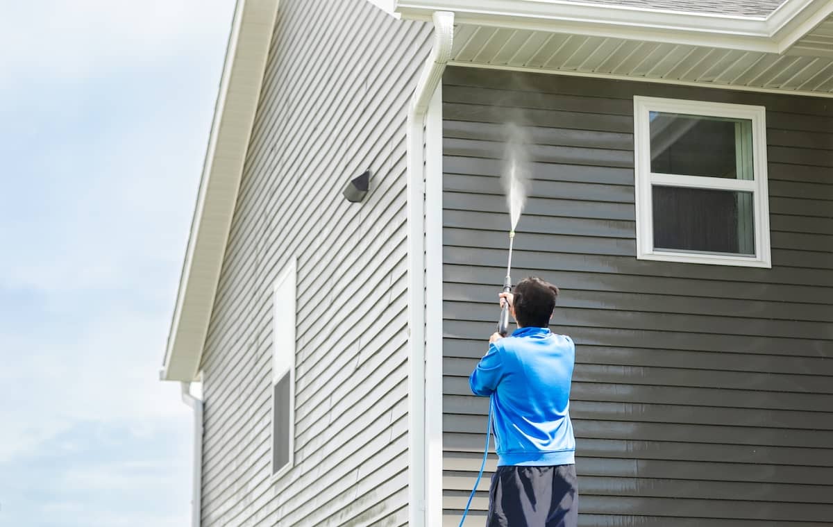 Man in blue jacket clean dusk and dirt from exterior siding and under roof by high-pressure nozzle spray with water soap cleaner