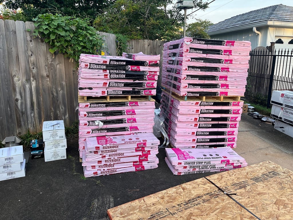 Stacks of pink and black roofing materials labeled "Duration" and "Ridge Vent" are piled on wooden pallets in an outdoor area next to a wooden fence and greenery. Some roofing boxes and planks lie nearby on the ground.