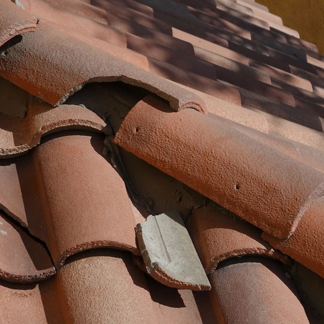 Close-up of red clay roof tiles with one piece visibly broken. The broken tile is located near the ridge, showing signs of damage and potential weathering. The surrounding tiles appear to be intact, casting shadows from sunlight. This scene illustrates typical The Woodlands roof damage.
