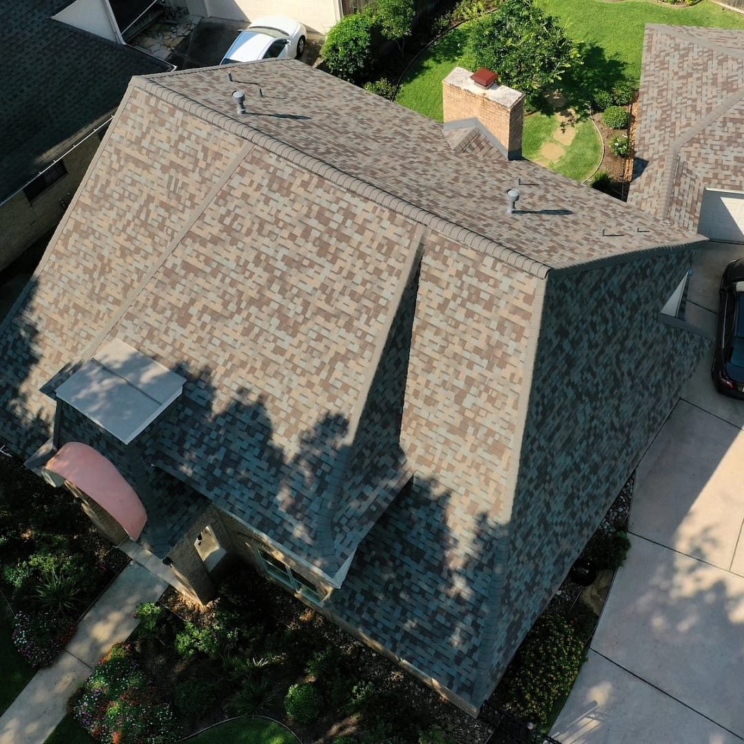 Aerial view of a residential home with a multi-gabled roof covered in shingles, part of a recent four corners roof replacement. The house is surrounded by a well-maintained garden, driveway, and neighboring houses. Shadows of nearby trees are cast on the new roof, and a car is parked in the driveway.