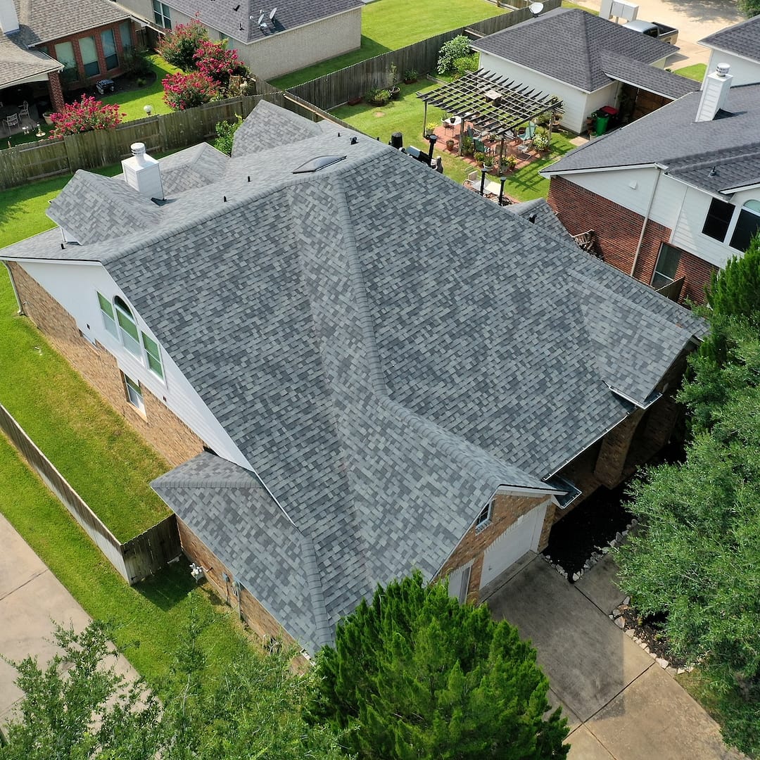 Aerial view of a suburban house in central northwest Houston with a new, grey shingle roof. The brick exterior home features an attached garage. Nearby houses, a wooden fence, a small backyard garden, and a well-maintained lawn are visible. Trees partially surround the property after its roof replacement.