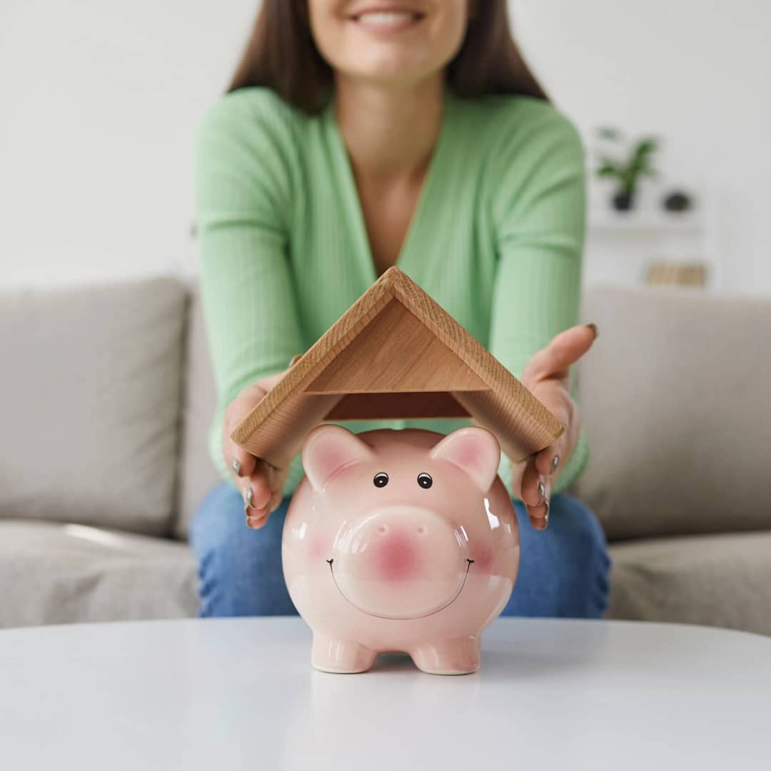 A person wearing a green sweater is sitting on a couch in the background, holding a small wooden roof above a smiling pink piggy bank placed on a white table in the foreground, symbolizing saving money or financial protection, reminiscent of Kingwood roof replacement efforts.