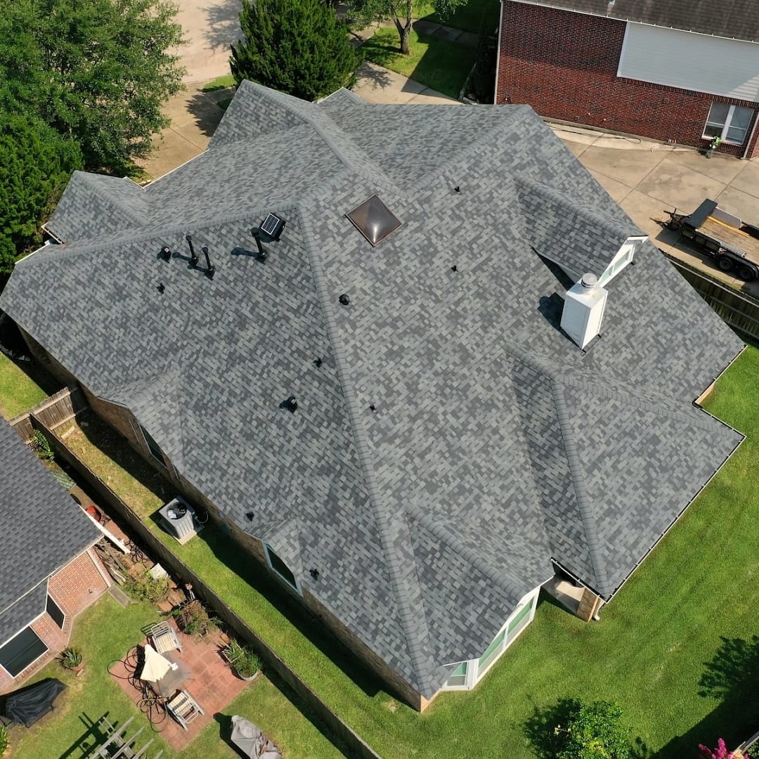 Aerial view of a gray-roofed house with multiple gables and a chimney. The roof features various skylights and roof vents, serviced by Four Corners Roof Repair. The property includes a patio area on the left side with garden furniture, located in a green, well-maintained backyard.