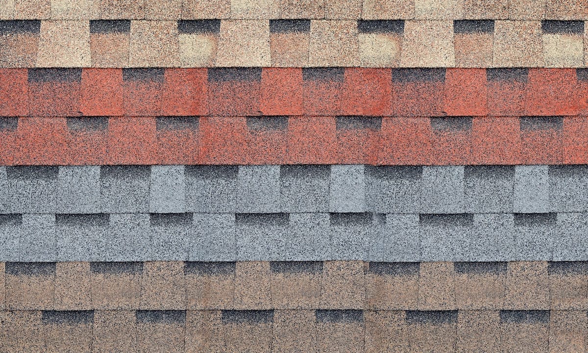 Close-up of a wall featuring multiple rows of different colored roof shingles, as typically seen in Houston roofing. The shingles are organized in horizontal layers, each layer showcasing a distinct color: beige on top, followed by red, grey, brown, and dark brown at the bottom.