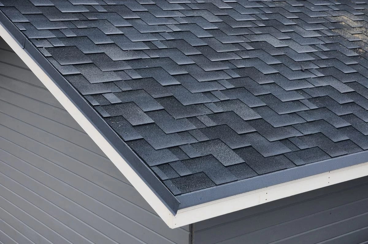 A close-up view of a house roof, showcasing dark gray asphalt shingles arranged in a staggered pattern. The roof edges are bordered with white trim, and the exterior siding of the house is painted in a complementary gray color. This Houston roof replacement highlights the clearly visible roof slope.