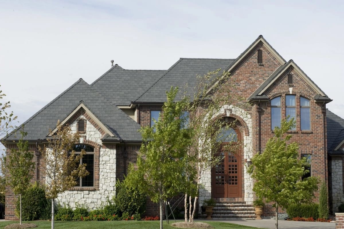 A large, two-story brick and stone house with tall arched windows and a wooden front door stands proudly. The home features a well-manicured lawn and several young trees. Recently benefiting from a Houston roof replacement, the steeply pitched roof gleams under the clear sky.