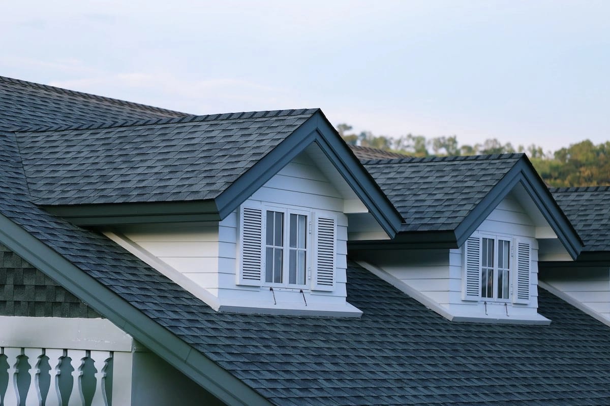 A close-up of a house roof with gray shingles, possibly slated for a Houston roof replacement. The roof features two dormer windows with white wooden shutters. The sky is clear and light blue, and some trees are visible in the background. A section of a white railing is visible on the lower left.