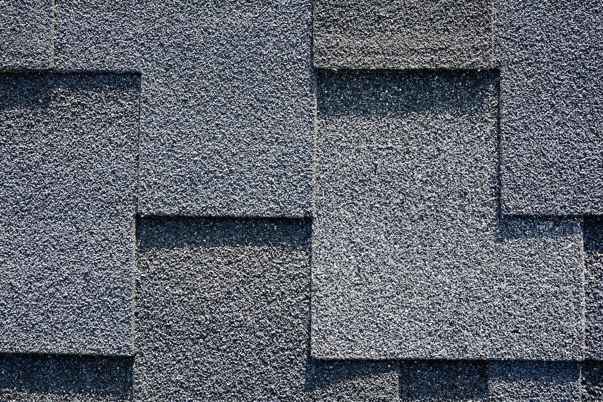 Close-up of a textured gray asphalt shingle roof with overlapping rectangular shingles. The surface has a rough, granular appearance typical of Houston roofing materials, arranged in a staggered pattern. Lighting highlights the texture and creates subtle shadows.