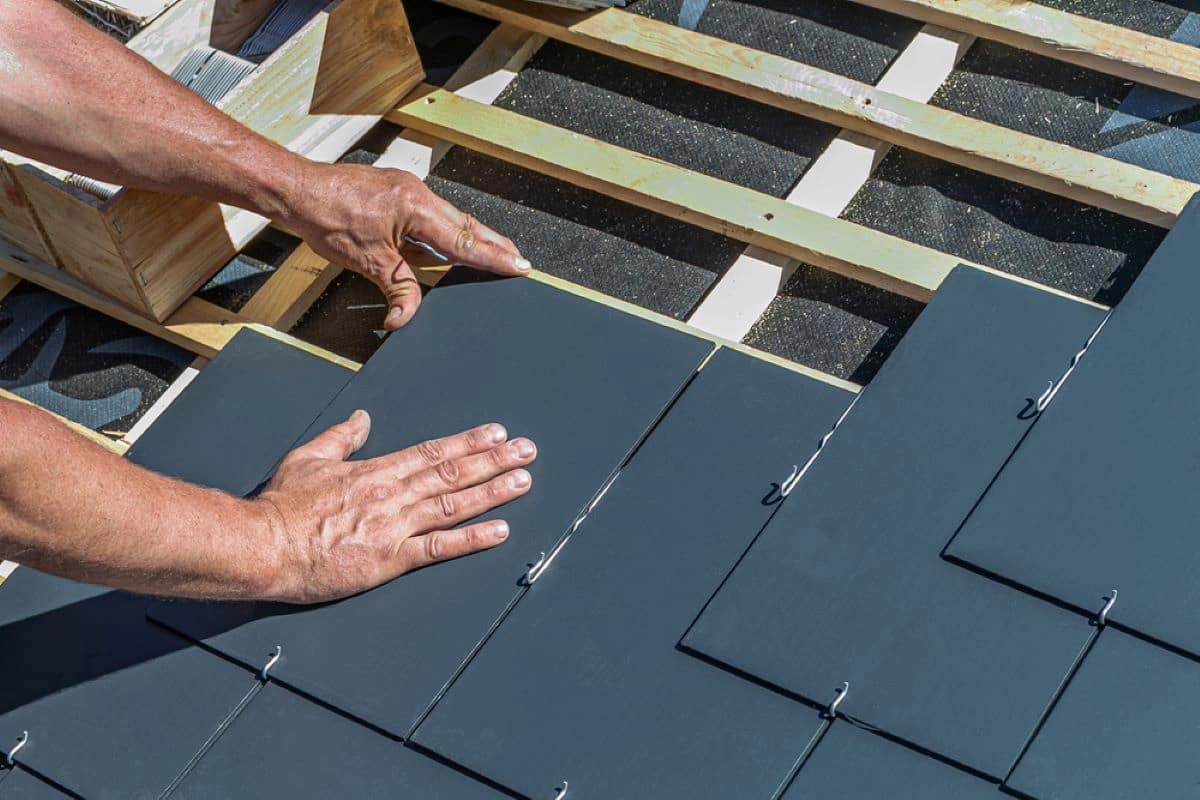 Close-up of a worker's hands installing slate roof tiles on a wooden frame. The tiles are aligned in neat rows, and the worker is positioning a tile, ensuring it fits securely. The background shows other construction materials and partially completed roofing by DaVinci Roofing.