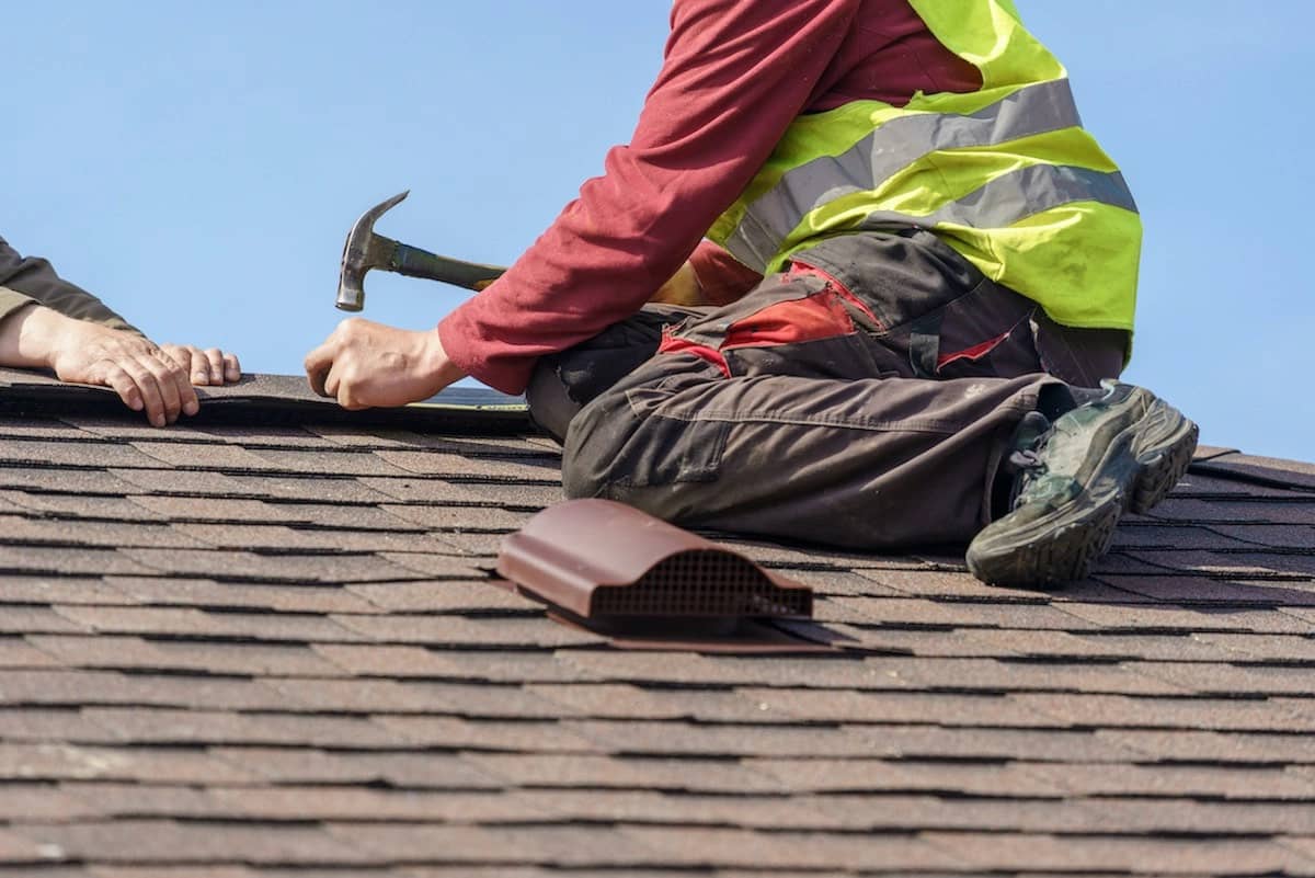 A construction worker in a high-visibility vest and red long-sleeve shirt is kneeling on a shingled roof, using a hammer. Another person’s arm is visible, assisting with the Houston roofing task. A vent lies on the roof surface nearby. The sky is clear above.