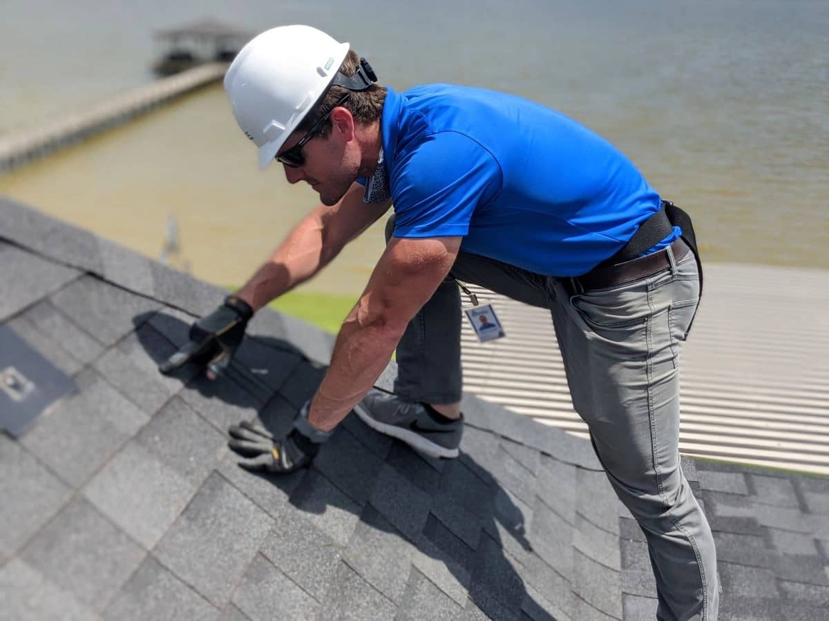 A man in a blue polo shirt, gray pants, and a white hard hat and safety harness is inspecting a shingled roof near a body of water. He is wearing gloves and sunglasses, closely examining the shingles as part of Houston roof repair services.
