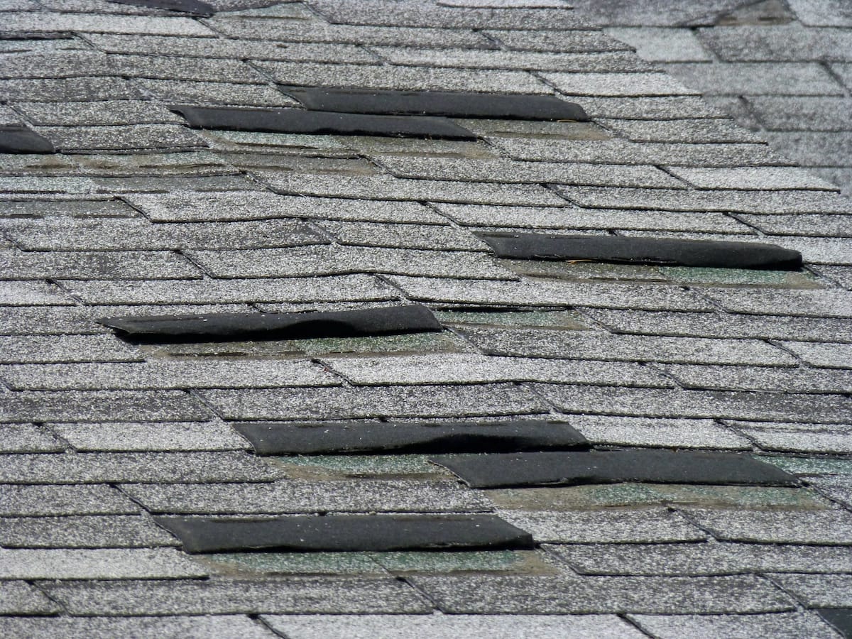 An image shows a close-up view of a shingle roof with several damaged and curled shingles. The roofing material appears aged and in need of repair, making it clear that a Houston roof replacement is necessary. The shingles have a gray color, with some showing wear and gaps.