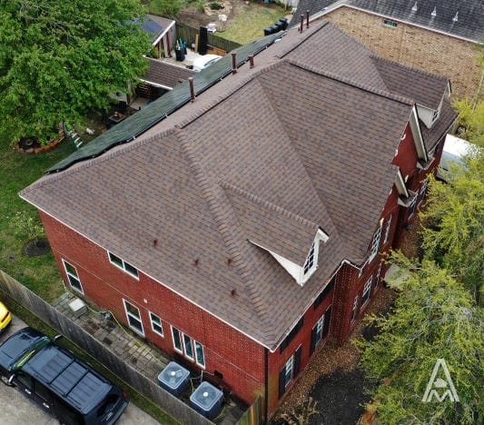 Aerial view of a multi-gabled, brown-roofed, two-story red brick house with several windows. The property includes a fenced backyard with a patio, large trees, and several parked vehicles, including a black SUV. The roof appears well-maintained but might benefit from some minor roof repair.