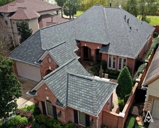 Aerial view of a large, two-story brick house with multiple gabled roofs and grey shingles, hinting at a recent roof replacement. The home features a three-car garage, well-manicured shrubs, and small trees around the property. Neighboring houses made of similar materials are visible on both sides.