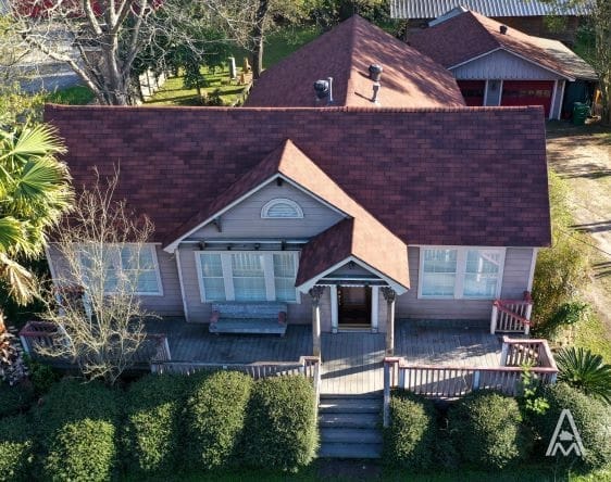 Aerial view of a quaint house with a large porch. The roof, which has recently undergone roof replacement, is reddish and the exterior walls are light-colored. The porch has a wooden railing and steps leading to the front door. The yard is surrounded by greenery, and a detached garage is visible in the background.