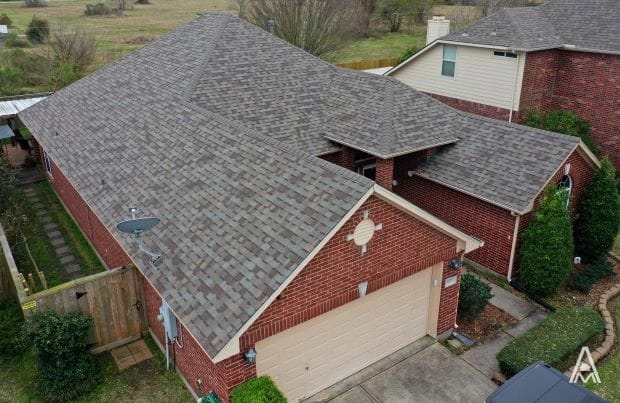 Aerial view of a red brick house with a grey shingled roof, hinting at a recent roof replacement, and a beige garage door. The house is surrounded by a wooden fence and a small, neatly kept lawn. Another house with similar design is visible in the background, and a satellite dish is attached to the fence.