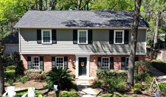 A two-story house with a red brick lower half and gray upper half situated in a landscaped yard, recently underwent a roof replacement. The house features black shutters, white trim, and a central black door with sidelights and a small porch cover. Tall trees surround the yard, providing shade.