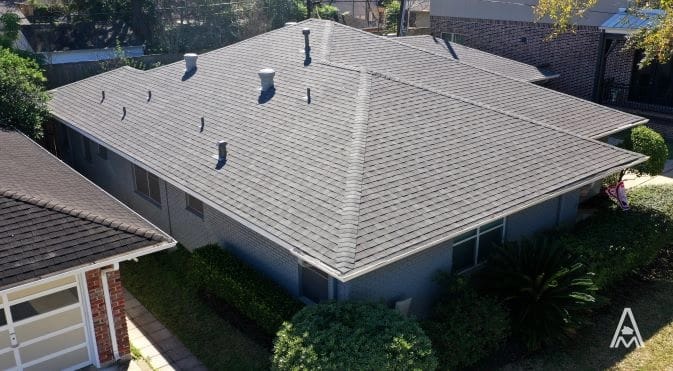 An aerial view of a single-story house with a grey shingled roof hints at the need for roof replacement. The surrounding area includes a green yard with bushes and trees, a neighboring building with a similar design, and a concrete pathway leading to the entrance.