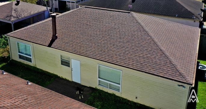 Aerial view of a single-story house with a brown hipped roof and beige exterior. The house features a white door, two windows, and a central chimney. The lawn is partially visible around the house. Signs of recent roof replacement are evident as the corner of another roof can be seen in the foreground.