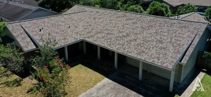 Aerial view of a single-story house with a gray shingle roof, indicating a recent roof replacement. The property features a concrete driveway, small shrubs, and trees surrounding the house. Roof vents are visible, and the house is set in a neighborhood with other homes partially visible in the background.