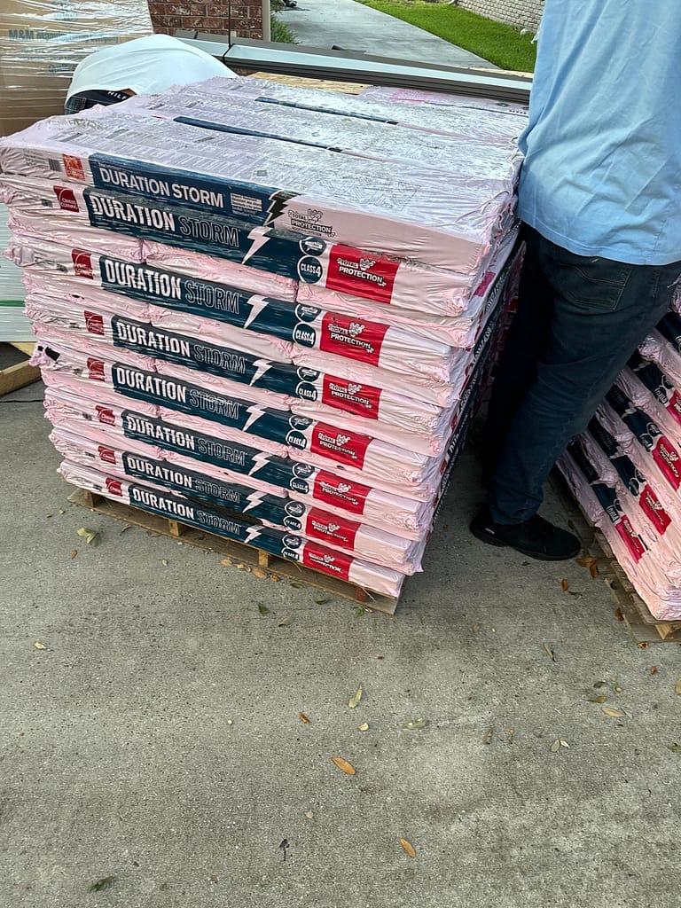 Stacks of Duration Storm roofing materials wrapped in pink packaging sit on a concrete surface. A person in blue jeans and a light blue shirt stands next to the stacks, with only their legs visible. Leaves are scattered on the ground.