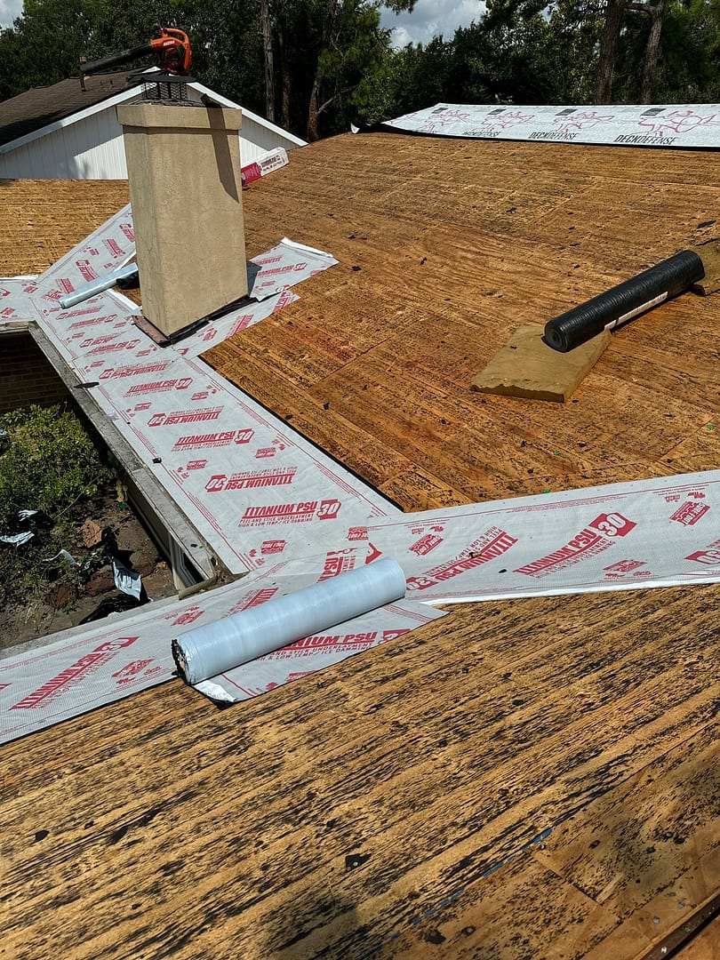 A partially renovated residential roof with wooden shingles and roofing underlayment in progress. A chimney protrudes through the roof. Nearby are tools and rolls of underlayment material. Trees and a neighboring roof are visible in the background.
