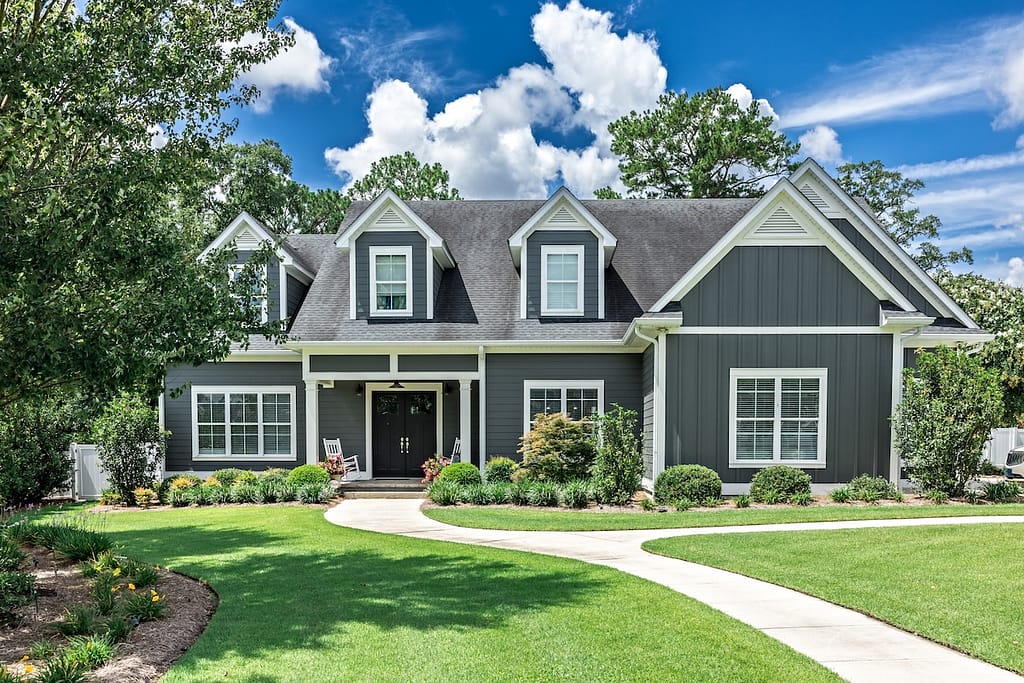 A large gray craftsman new construction house with a landscaped yard and leading pathway sidewalk on a sunny day with blue skies and clouds.