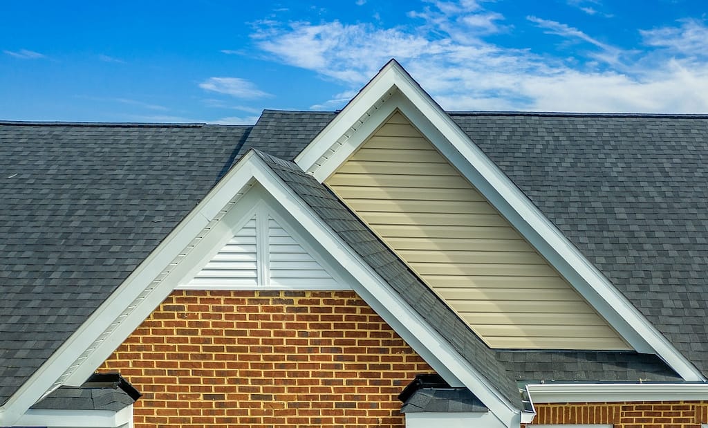 Double gable with beige horizontal vinyl lap and red brick facade siding, with triangle shape white attic vent on a pitched roof attic at an American single family home neighborhood USA