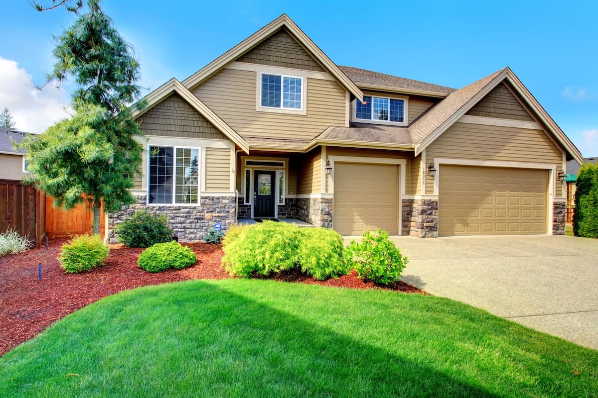 Clapboard siding house with stone trim. View of entrance porch, beautiful flower bed with green lawn, bushes and fir tree