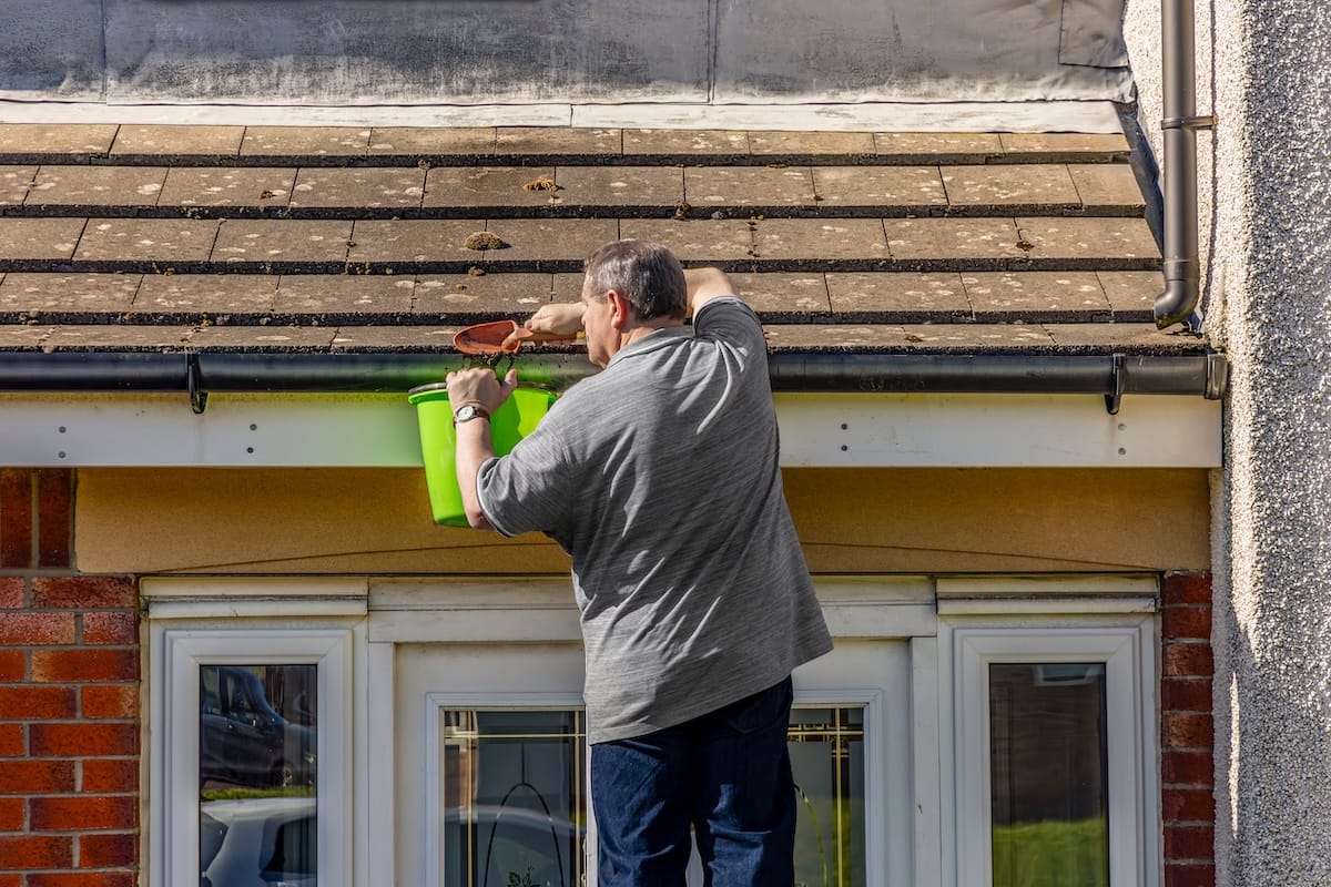 Man on ladder cleaning leaves and moss from house roof guttering above the front door of a house. He uses a trowel to remove the debris and places it in a green bucket that he holds in his left hand. This is ideal to illustrate for home maintenance, gutter cleaning, DIY projects, and rooftop safety content.