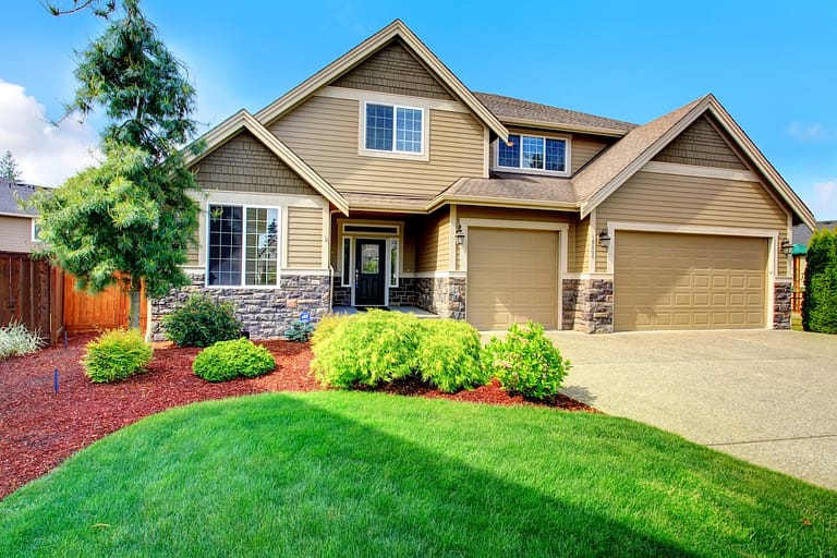 Clapboard siding house with stone trim. View of entrance porch, beautiful flower bed with green lawn, bushes and fir tree