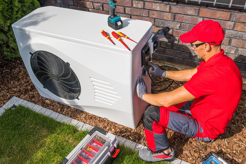 how much to replace a heating and air conditioning system Professional Middle Aged HVAC Technician in Red Uniform Repairing Modern Heat Pump Unit. House Heating and Cooling System Theme.