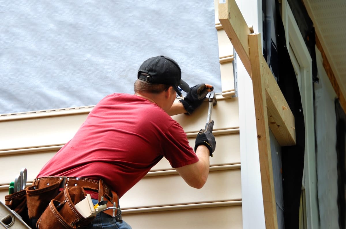 Worker installs siding to a home. He is holding a hammer and wearing a tool belt.