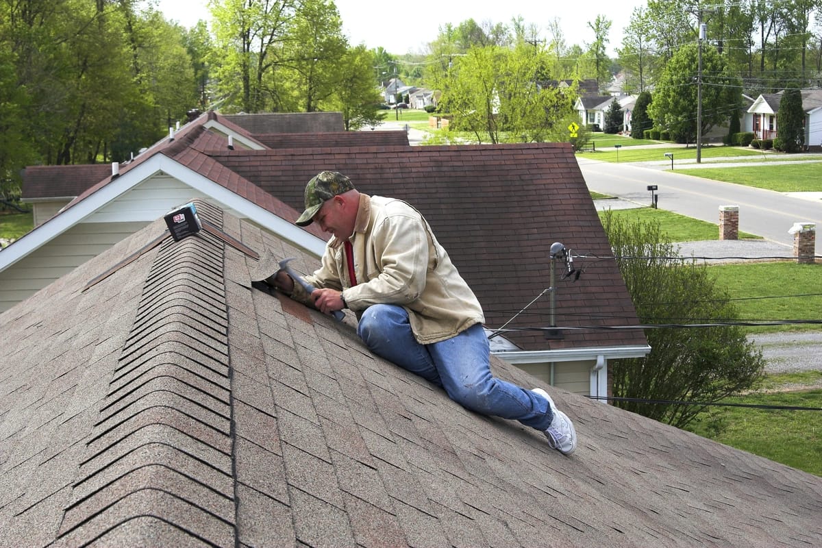 A person wearing a cap and casual clothes is carefully repairing shingles on a steep, sloped roof in a residential area. The neighborhood features houses with trees and a road in the background, all under a clear sky.