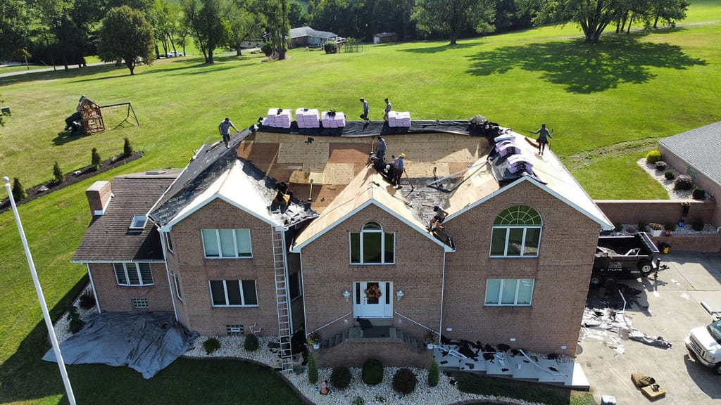 Aerial view of a two-story brick house with roofers replacing the shingles. The old shingles are being removed, and new underlayment is being installed. Supplies and bundles of shingles are stacked on the unfinished roof. Surrounding trees and lawn are visible.
