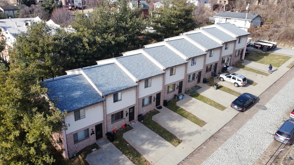 Aerial view of a row of townhouses with blue roofs and brick facades, lined along a paved driveway. Cars are parked in front of each unit, with trees and a few people visible around the area.